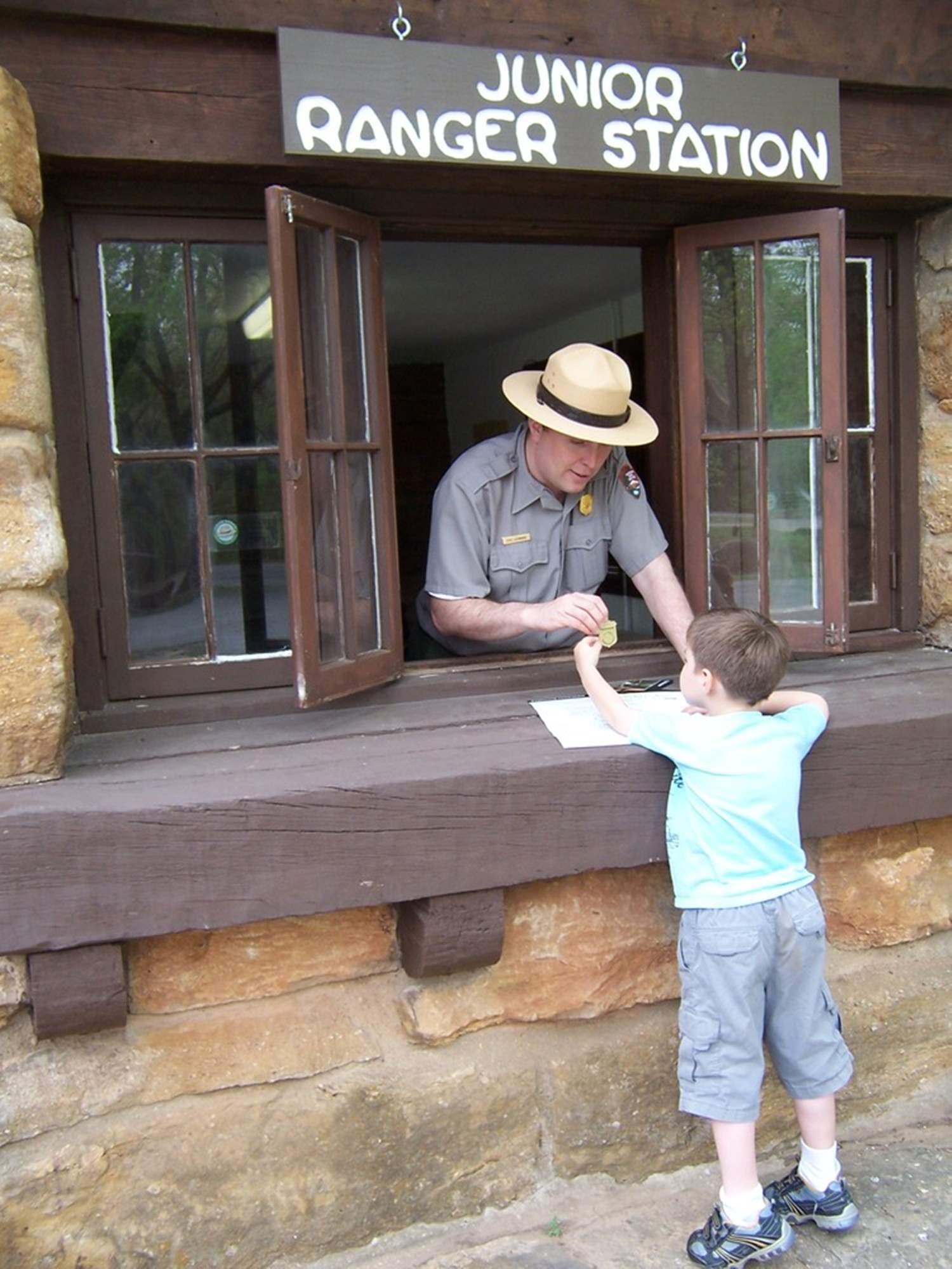 Man in broad brim hat and uniform with shield-shaped badge on his shirt leans out the Junior Ranger Station window to hand a boy a shield shaped badge.